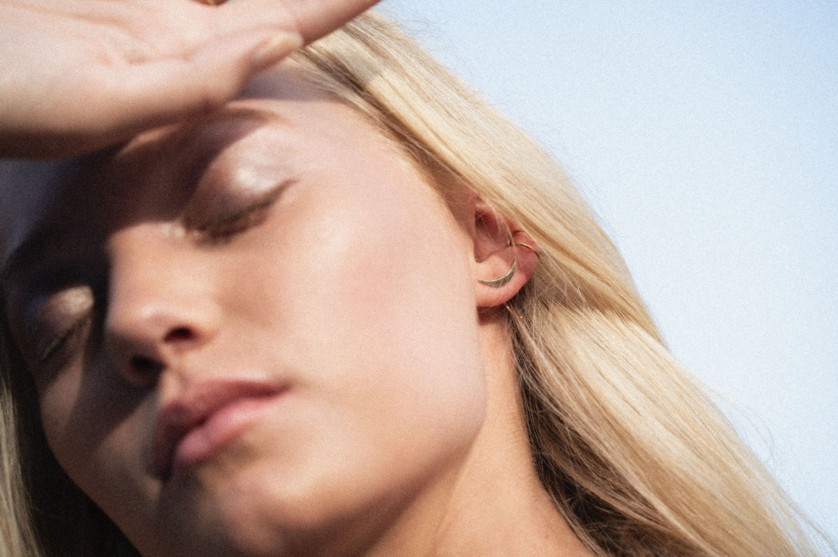 Close-up of a blonde woman outdoors with her hand over her eyes, wearing a small crescent moon earring on her ear.